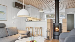 Una sala de estar moderna con sofá, una mesa redonda con frutas, chimenea y una zona de cocina al fondo.
