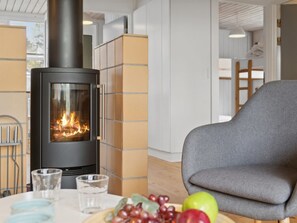 Un interior moderno con un estufa de leña, un sillón gris y una mesa con vasos y fruta.
