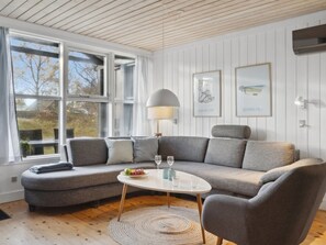 Una sala de estar moderna con un sofá modular gris, una mesa de centro redonda y una lámpara. Hay dos cuadros enmarcados en la pared, y la habitación tiene ventanas amplias con cortinas.