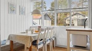 Un comedor con una mesa dispuesta para cuatro, sillas, una lámpara y vistas a una casa a través de amplios ventanales.