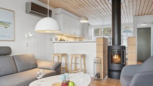 A modern living room with a sofa, a round table with fruit and glasses, a fireplace, and a kitchen area with stools.