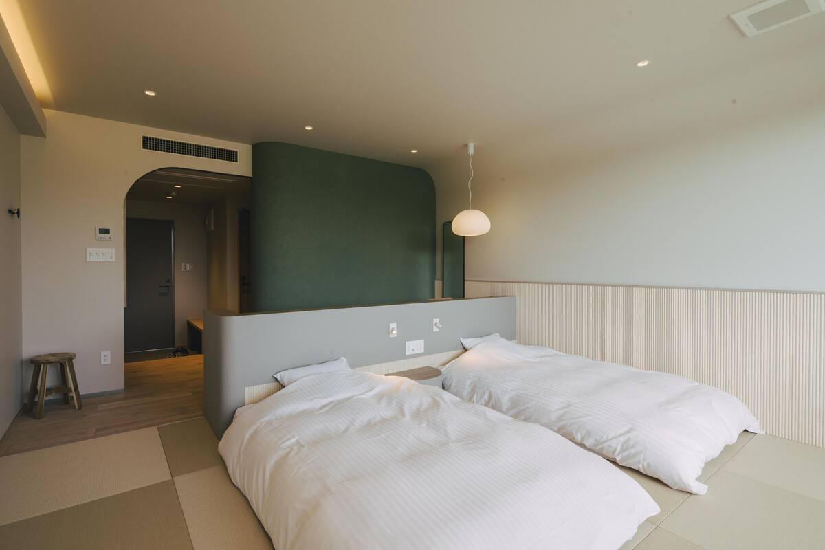 A modern hotel room with two beds, a small stool, and a green accent wall.