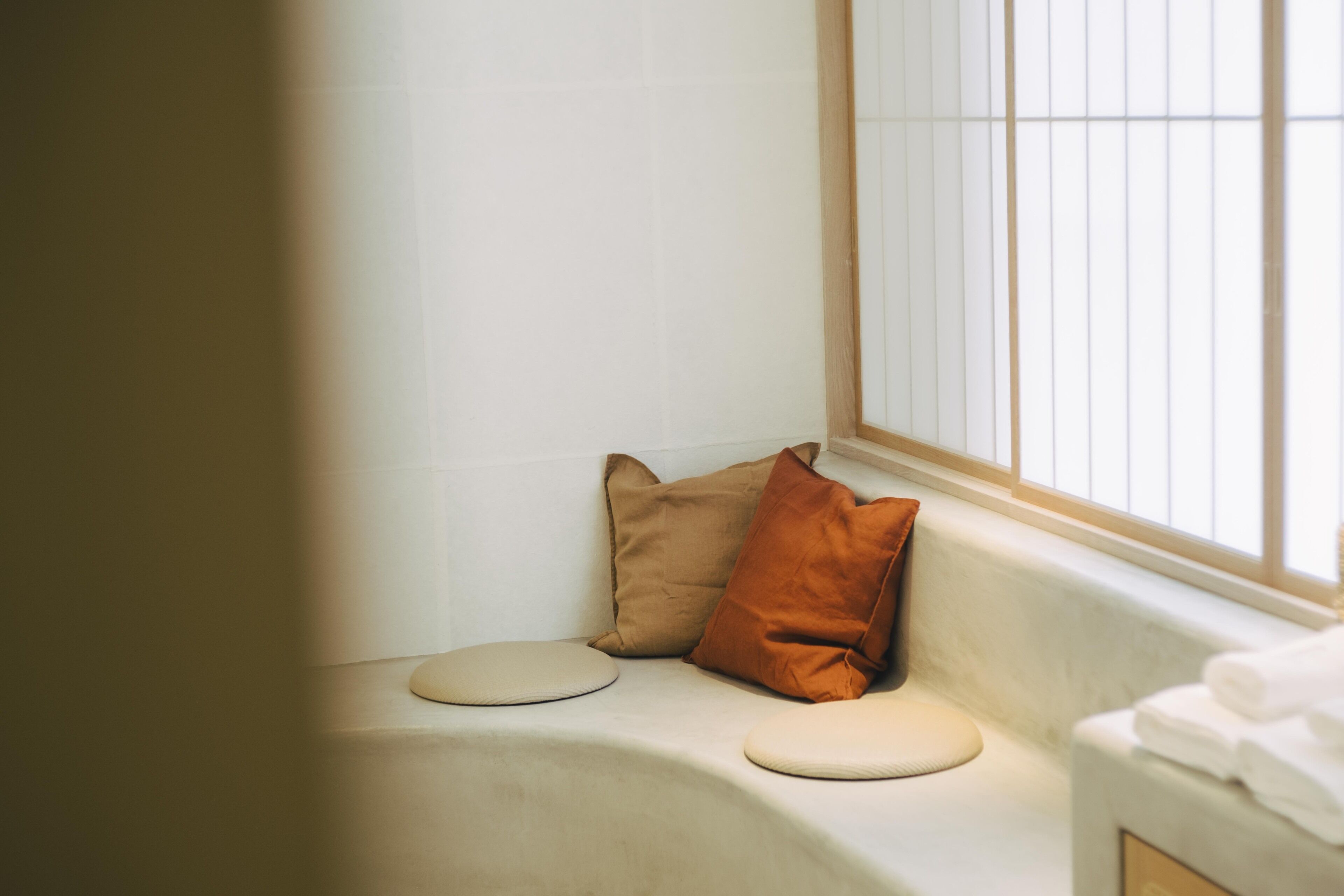 Een hoek van een kamer met een bankje, een raam met luiken en een handdoekhouder.