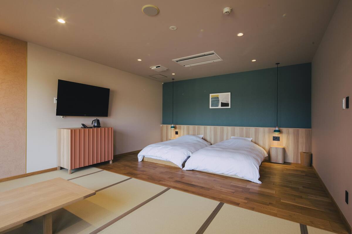 A modern hotel room with a wooden floor, a flat-screen TV mounted on the wall, a bedside table with a lamp, and a small cabinet.