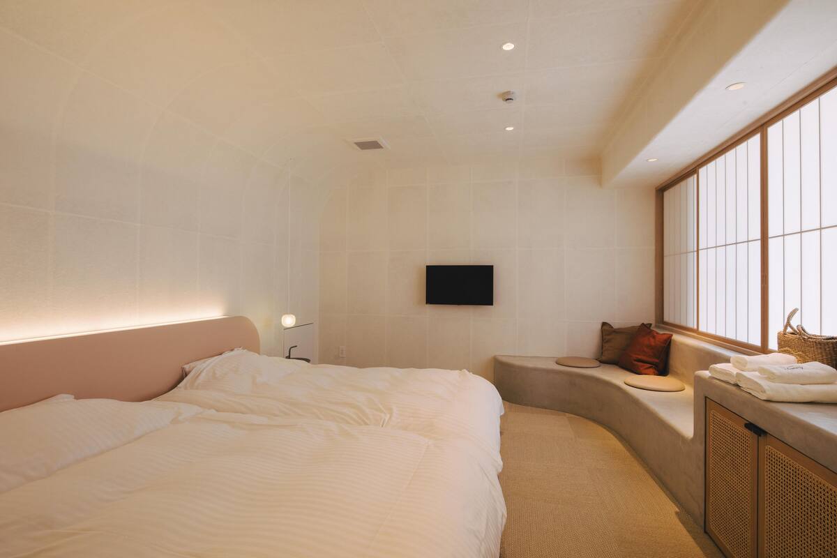 A modern hotel room with a large bed, a flat-screen TV mounted on the wall, a small seating area with cushions, and a large window providing natural light.