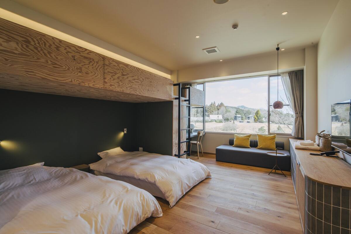 A modern bedroom with two beds, a desk, and a large window offering a view of the outdoors.