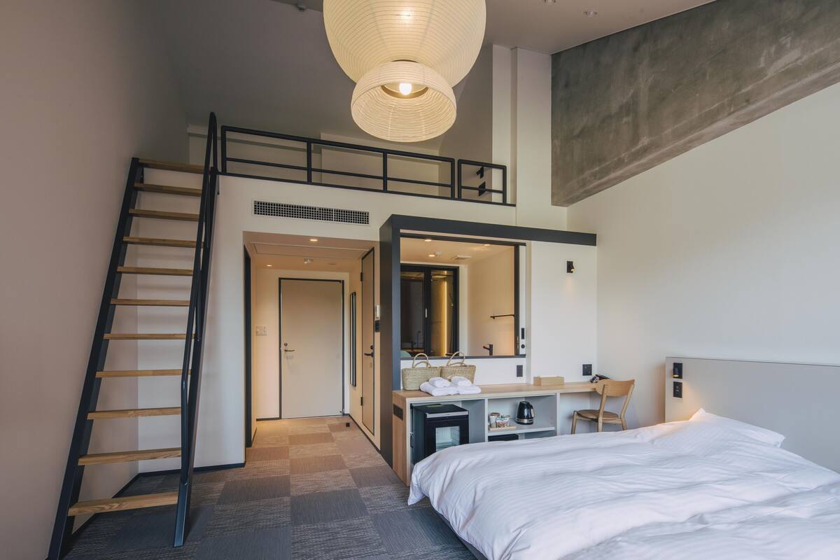 A modern hotel room with a lofted sleeping area, a desk with a chair, a large bed, and a staircase.