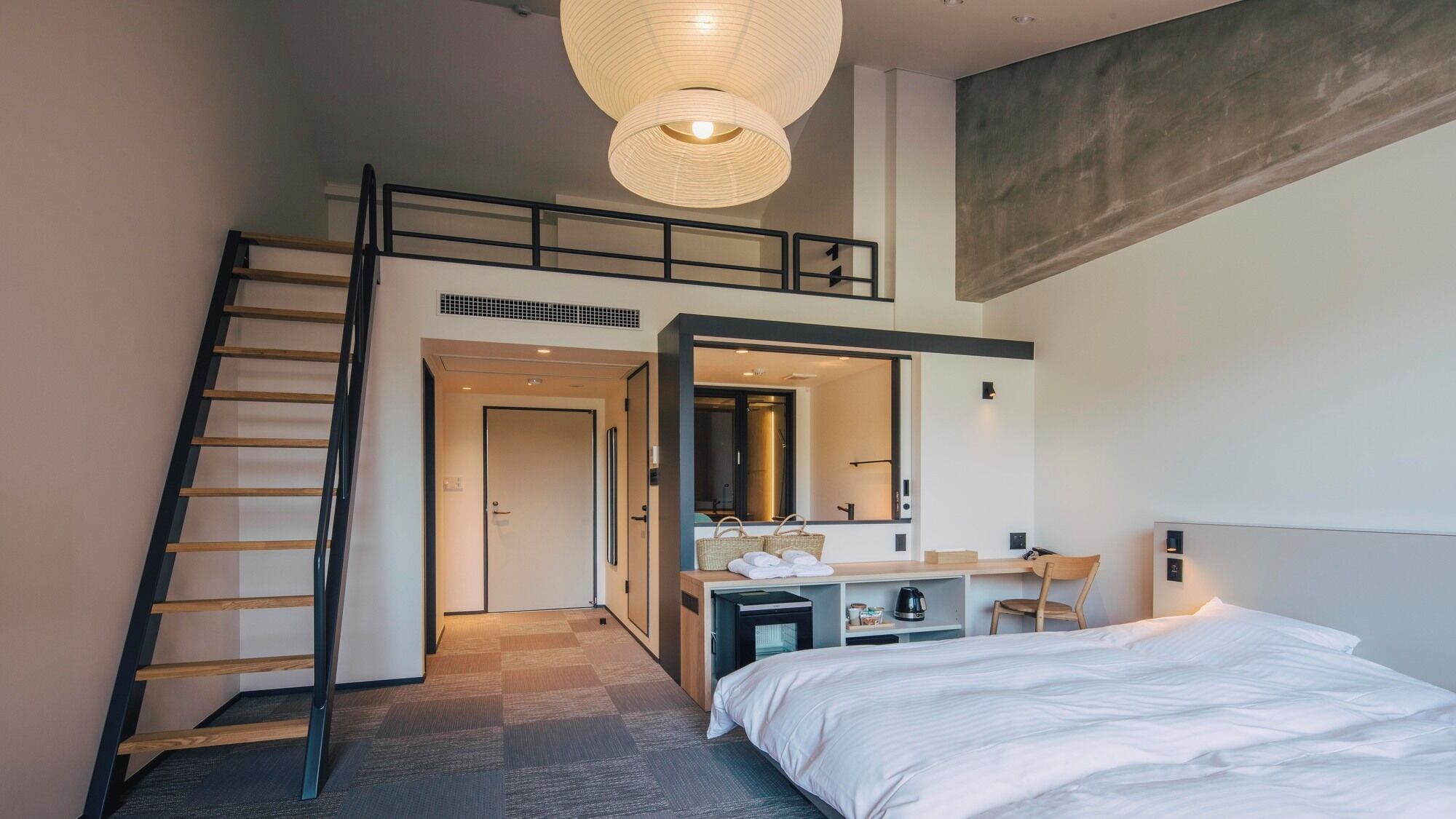 Habitación de hotel moderna con cama tipo loft, una escalera, un baño con un espejo grande y una lámpara colgante.