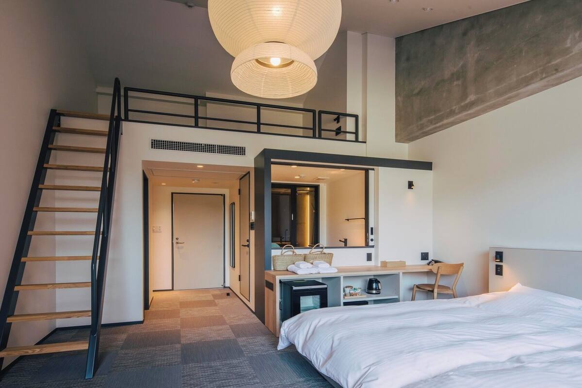 A modern hotel room with a loft bed, a staircase, a bathroom with a large mirror, and a hanging light fixture.