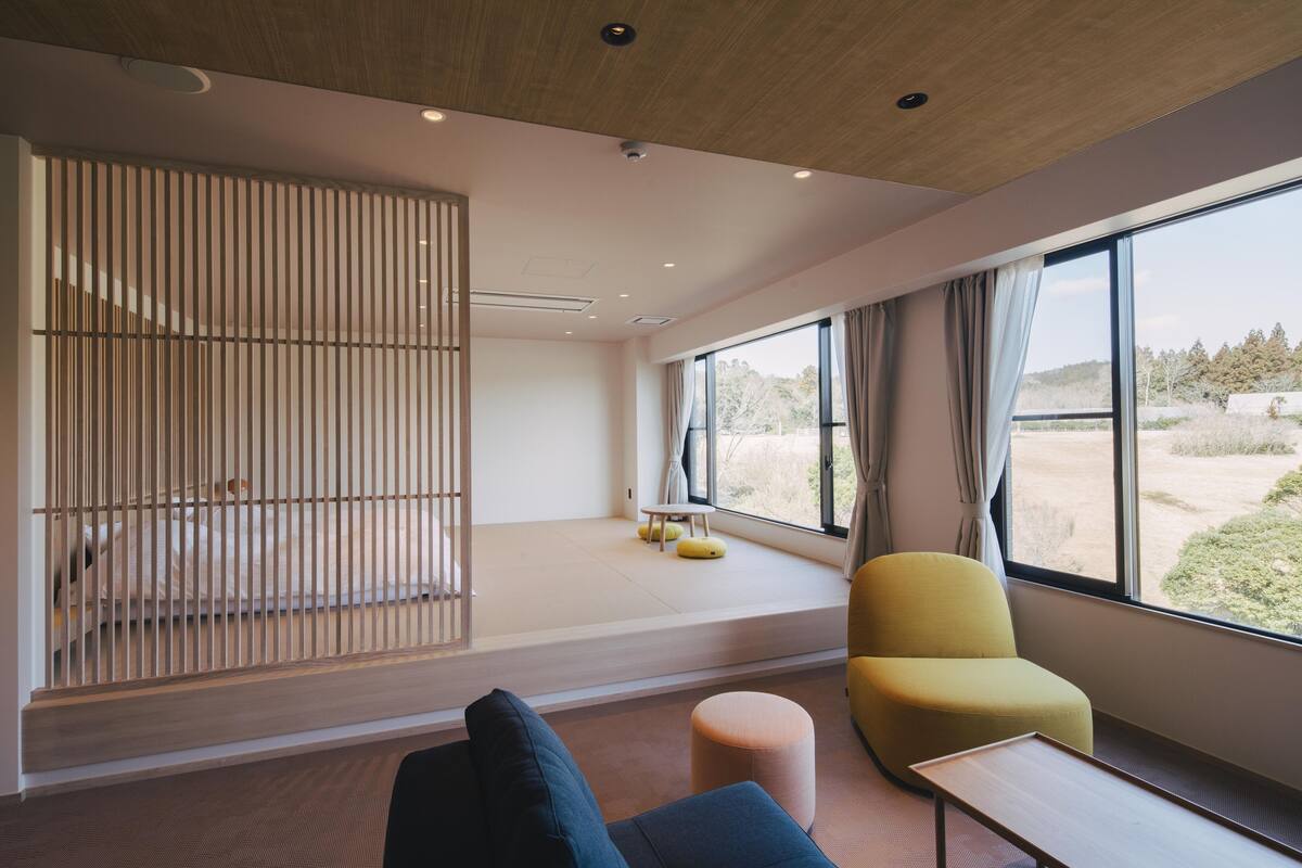 A modern room with a large window, a yellow chair, a small table, and a bed with a wooden slat partition.