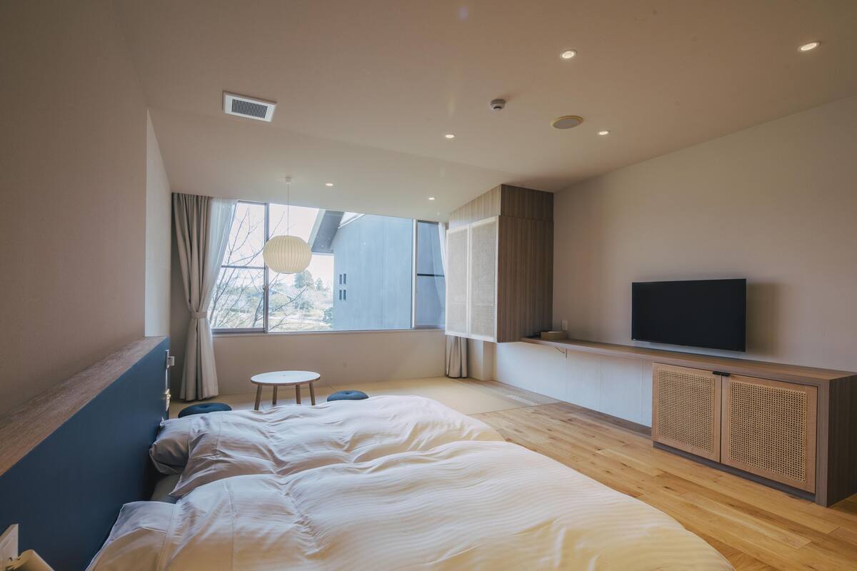 A modern bedroom with a large bed, a small round table, a TV mounted on the wall, and a window with curtains.