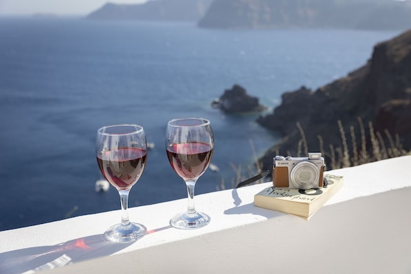 Two wine glasses on a white surface with a camera and a book beside them, overlooking a coastal view. - Kyrkos Residency (Santorini)