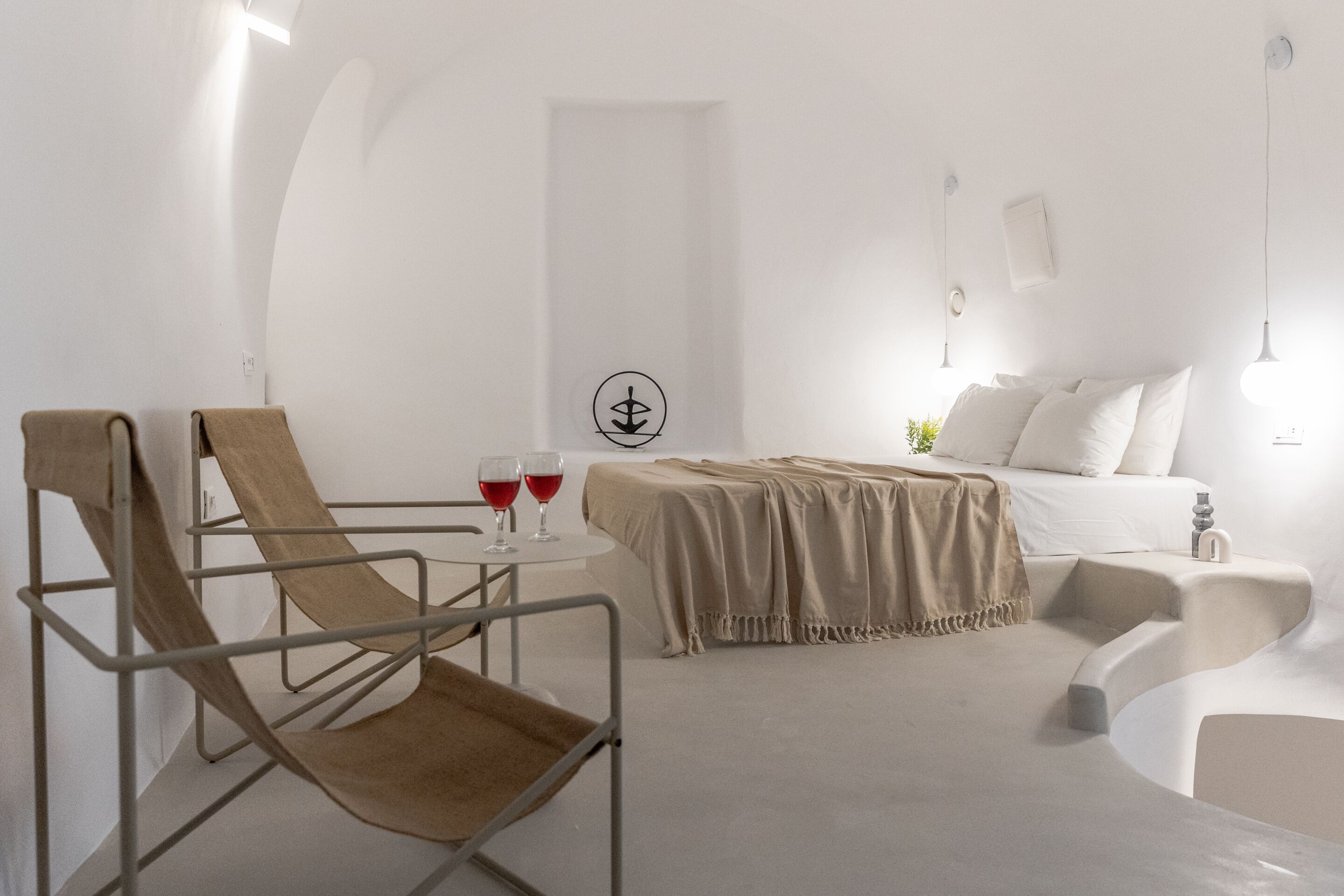 A minimalist room with a bed, a reclining chair, and a small table with two glasses of red wine.