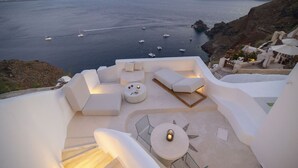 Eine Terrasse mit weißer Möbel, einem runden Tisch und Blick auf das Meer mit Booten.
