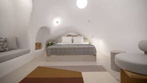 Ein modernes, minimalistisches Schlafzimmer mit gewölbter Decke, einem Bett mit gemusterten Betttextilien, einem grauen Sofa und einem runden Tisch.