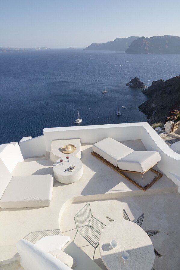 A sunlit outdoor seating area with white furniture and a view of the sea. - Kyrkos Residency (Santorini)