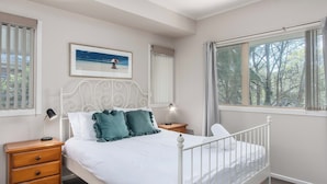 A bedroom with a white bed, a nightstand with a lamp, a framed picture on the wall, and a window with a view of trees. - Hastings Cove 2 Bedroom Upstairs Estuary Water View Apartment (Tweed)