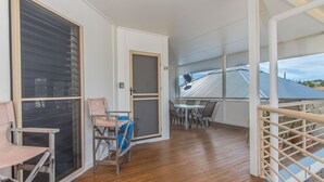 Un espacio al aire libre cubierto con piso de madera, una mesa con sillas, una puerta y vista a una casa con techo inclinado.
