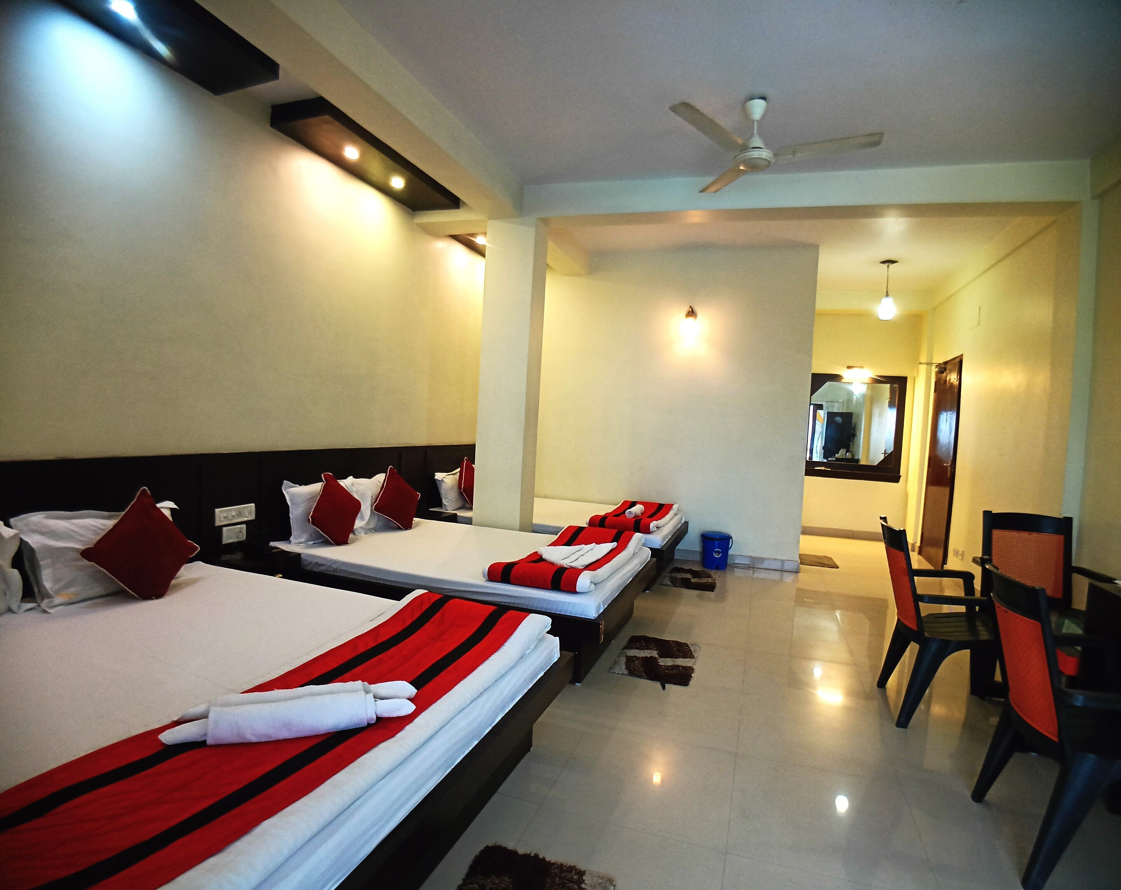 A hotel room with three beds, each with red and white bedding, a ceiling fan, and a small table with chairs.