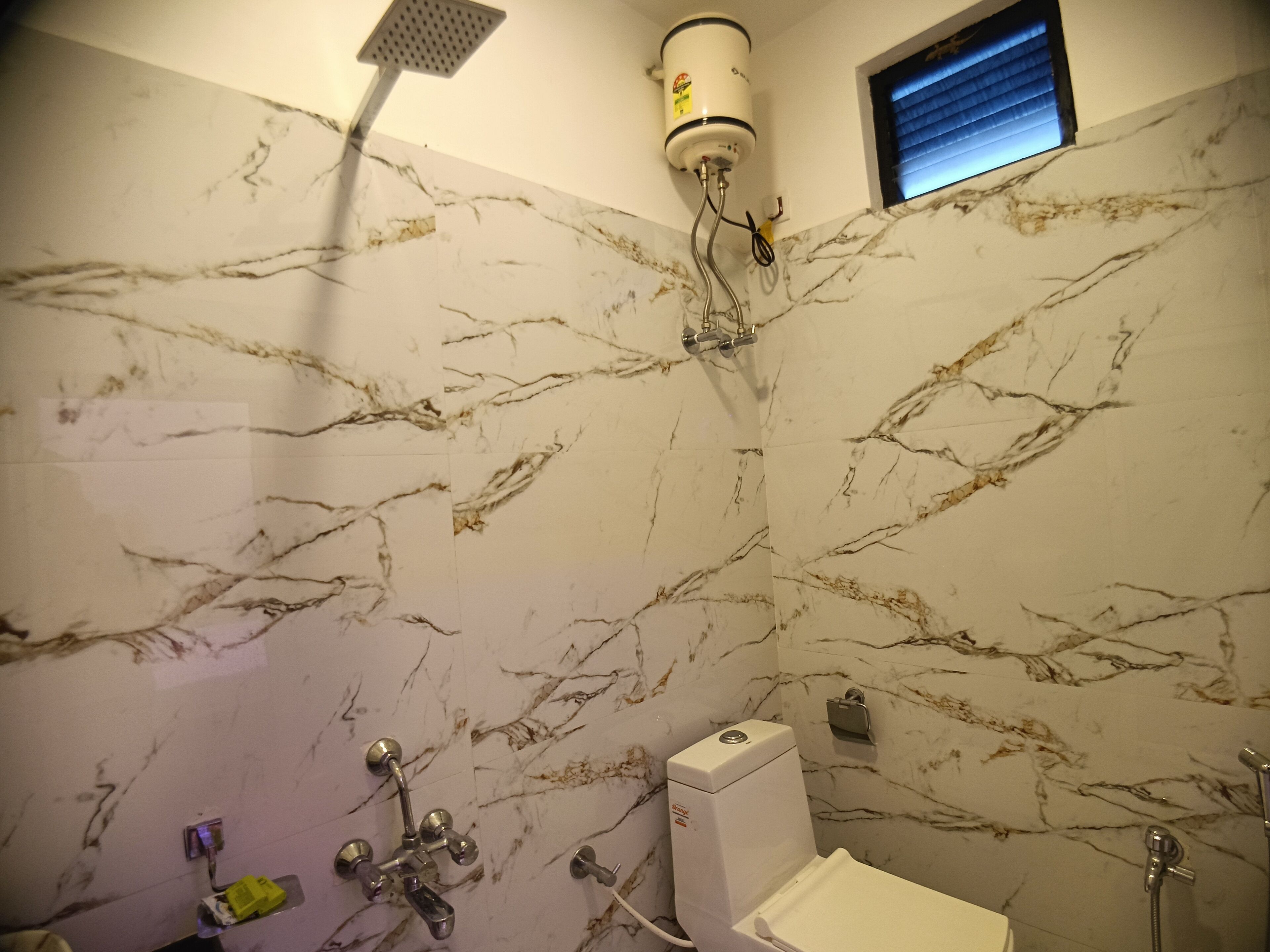 A bathroom with a marble wall, a handheld showerhead, a built-in toilet, and a window with blinds.