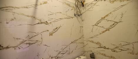 A bathroom with a marble wall, a handheld showerhead, a built-in toilet, and a window with blinds.