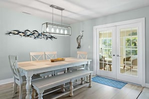 A dining area with a light blue table, white chairs, a bench, a modern chandelier, and a glass door leading outside.