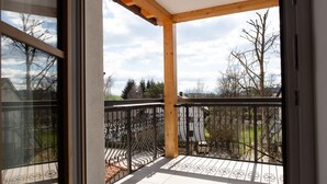 Un balcone con struttura in legno e ringhiera in ferro battuto nero, affacciato su una zona residenziale con alberi e un'auto parcheggiata.