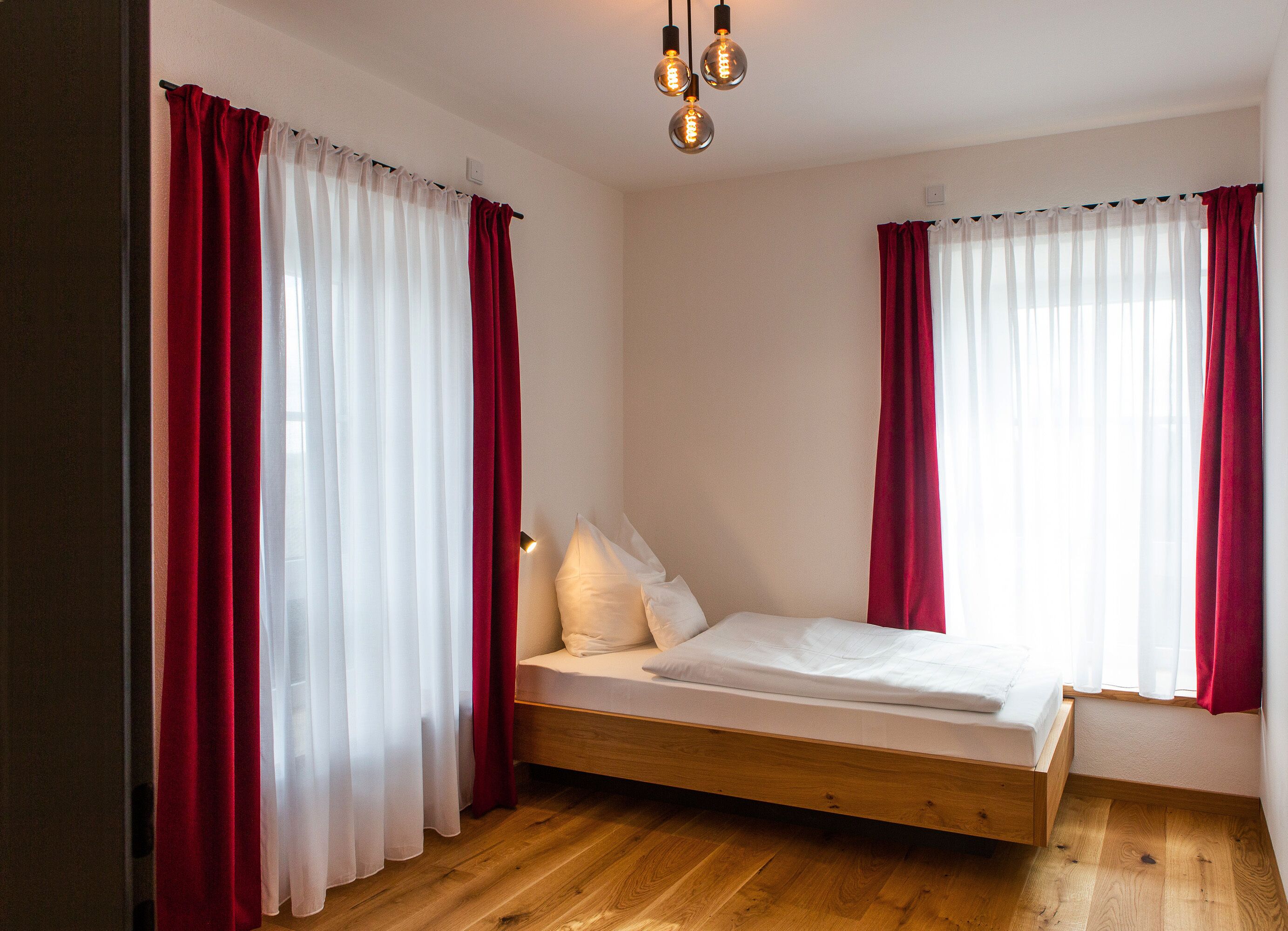 Ein Schlafzimmer mit einem hölzernen Bett, weißer Bettwäsche, roten Vorhängen und einer modernen Leuchte.