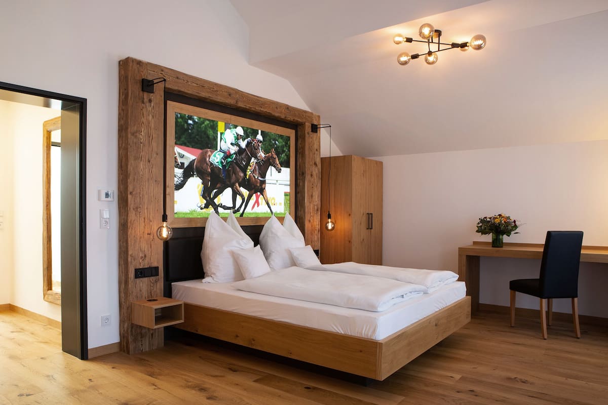 Une chambre moderne avec un cadre de lit en bois, un téléviseur fixé au mur diffusant une course de chevaux, un bureau avec une chaise et un luminaire suspendu.