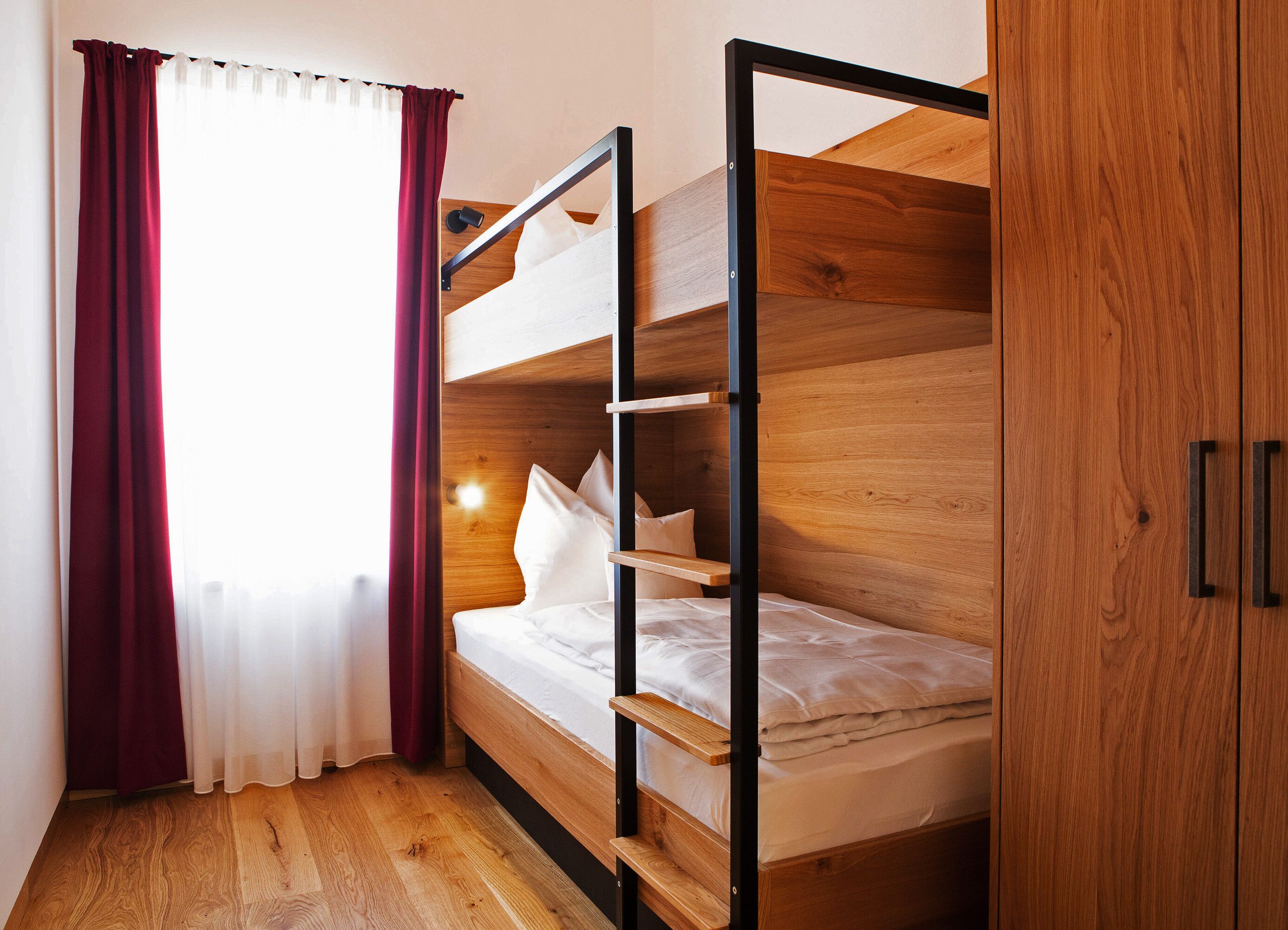 A bunk bed with a wooden frame and a single bed below. There is a window with white curtains and a wall-mounted light.