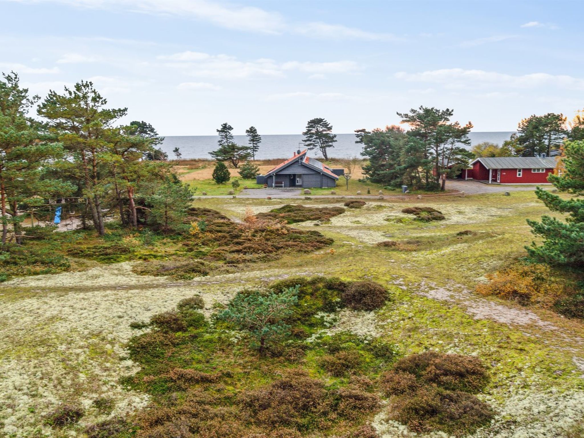 A remote cabin with a red exterior, surrounded by a natural landscape with sparse vegetation and a body of water in the background.