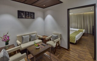Habitación de hotel con sofá, sillas, una mesita con fruta y un control remoto, y una cama con una manta verde.