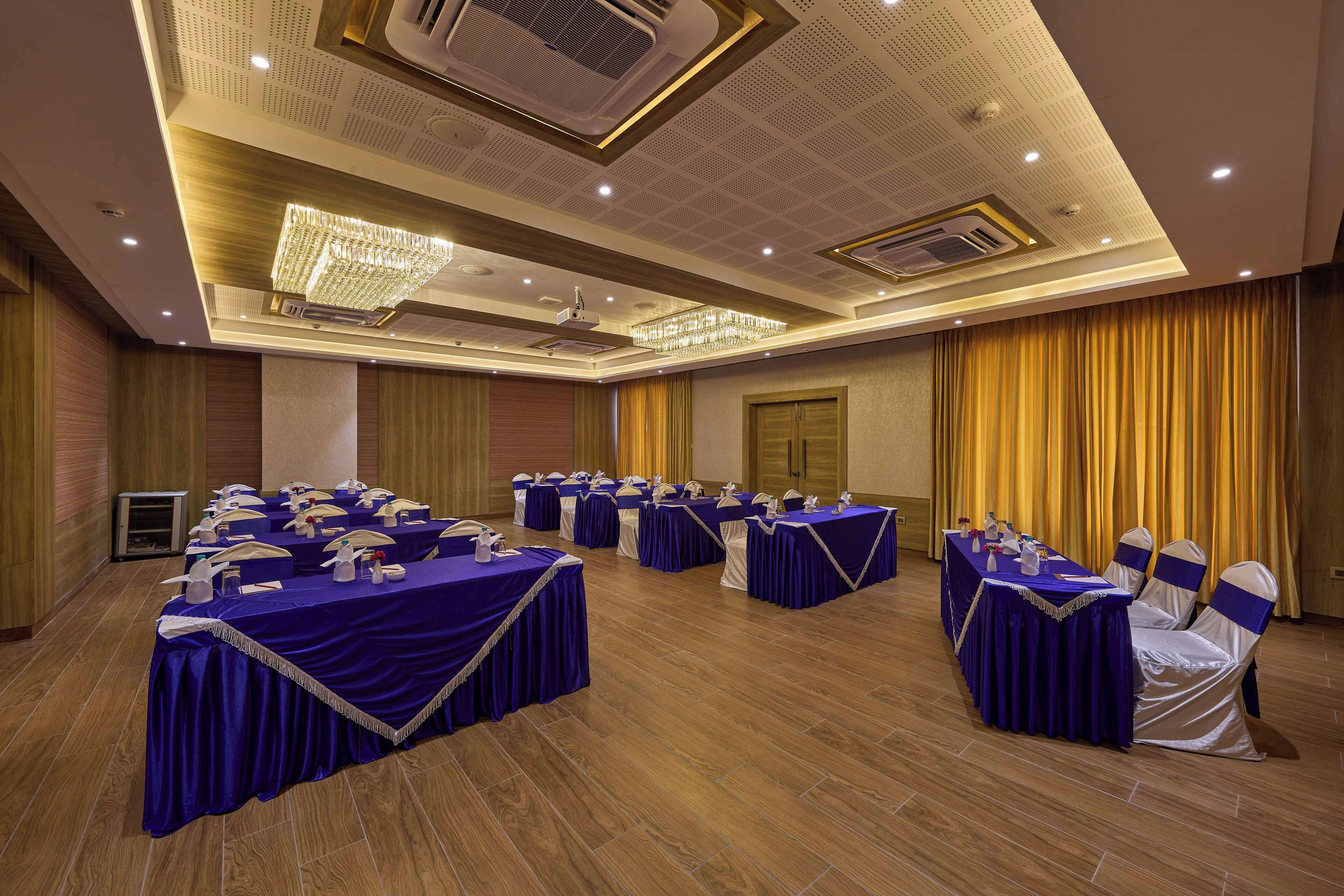 A conference room with multiple tables, each covered with a blue tablecloth and white chairs with blue sashes. The room features a wooden floor, a chandelier, and a ceiling with recessed lighting and air conditioning vents.