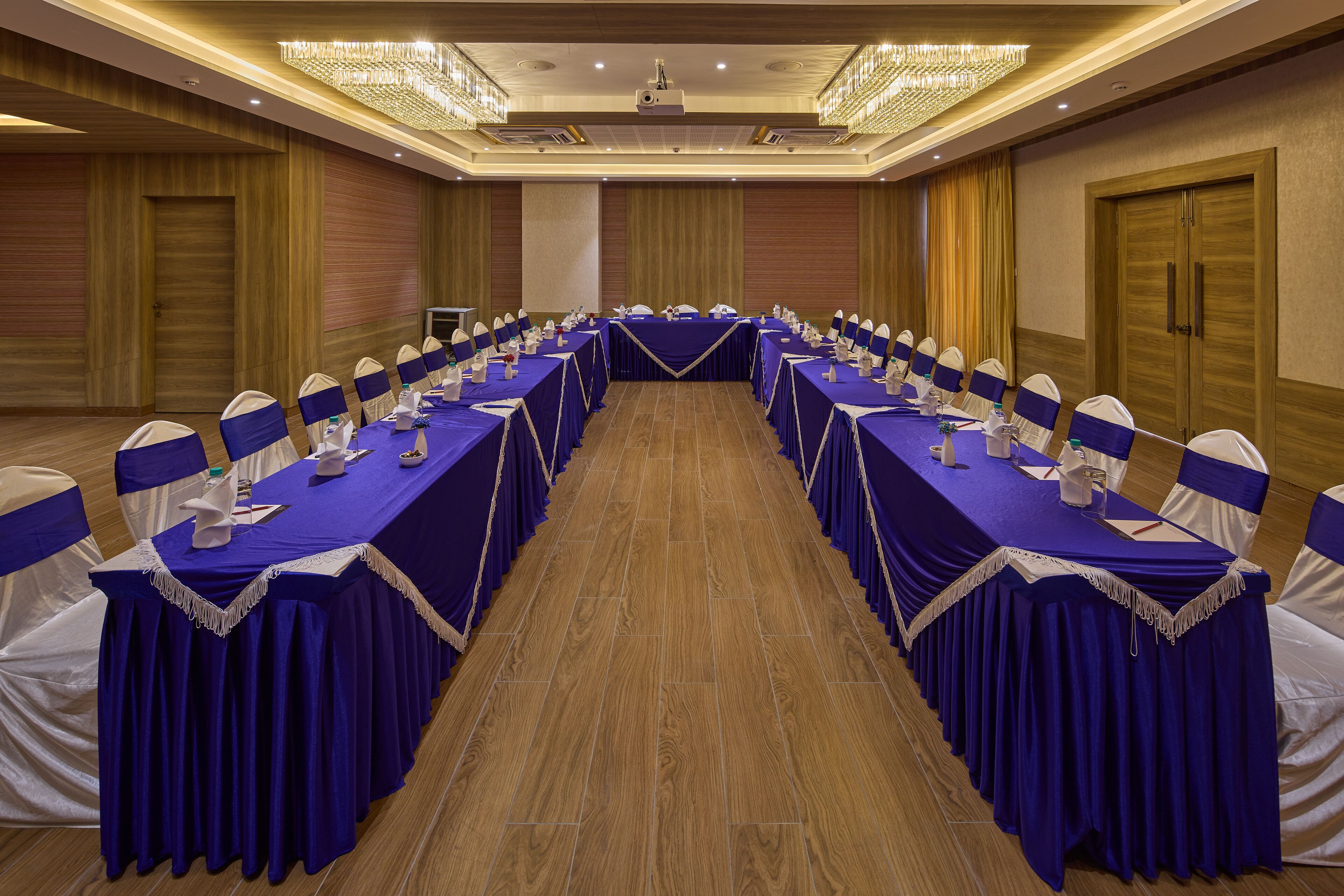 Un salón de conferencias con largas mesas, manteles azules y sillas blancas adornadas con cintas azules.