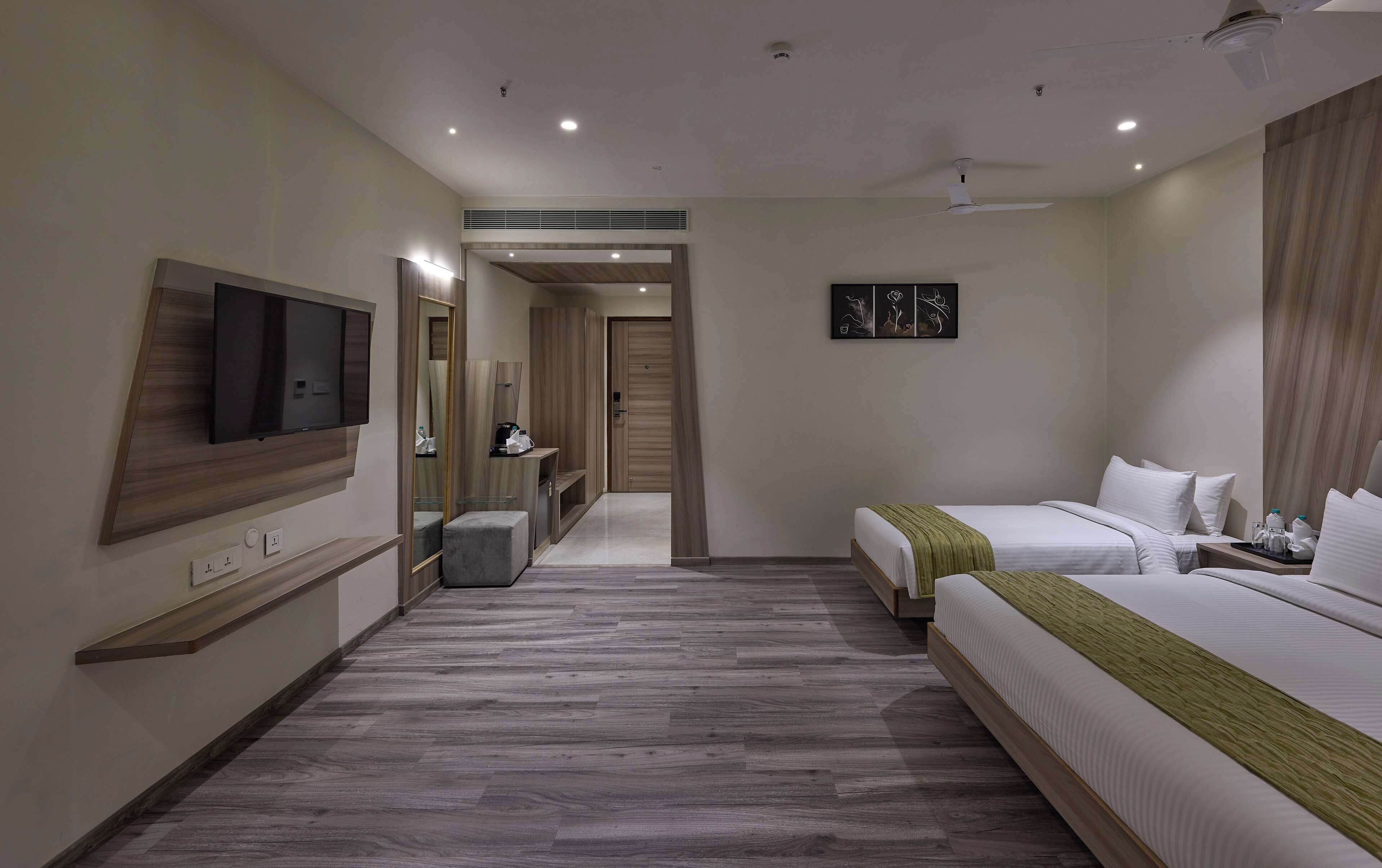 Une chambre d’hôtel moderne avec un grand lit, un lit plus petit, un téléviseur fixé au mur, un miroir et un couloir menant à une autre chambre.