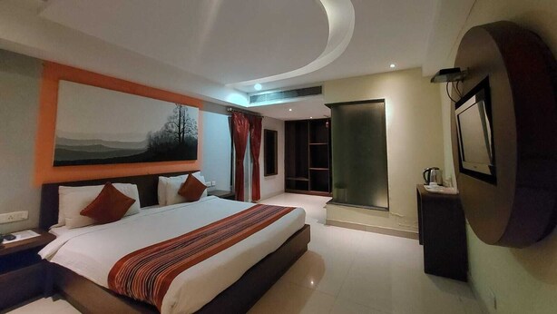 A modern hotel room with a large bed, a flat-screen TV, a wardrobe, and a bathroom with a glass partition. - Orbit Hotel Bagdogra (Siliguri)