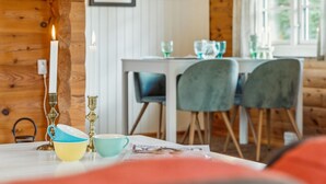 Ein gemütliches Zimmer mit Holzwänden, einem Tisch mit Kerze, einer Lampe und einer Zeitung. An der Wand befinden sich gerahmte Bilder und ein Fenster mit Blick.