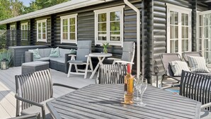 Eine Blockhütte mit einer Terrassenfläche, die Gartenmöbel, einen Tisch mit Flasche und Gläsern sowie einen großen Sonnenschirm zeigt.