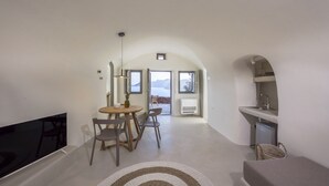 A modern interior with a flat-screen TV, a round dining table with chairs, a small kitchen area with a sink and a refrigerator, and large windows offering a view of mountains. - Oia Waves by SV (Santorini)
