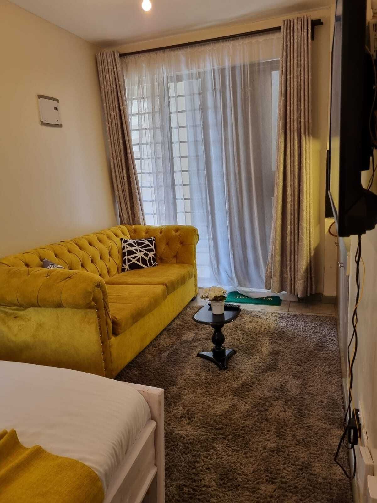 A room with a yellow sofa, a small table with a vase, a bed, and a window with curtains.