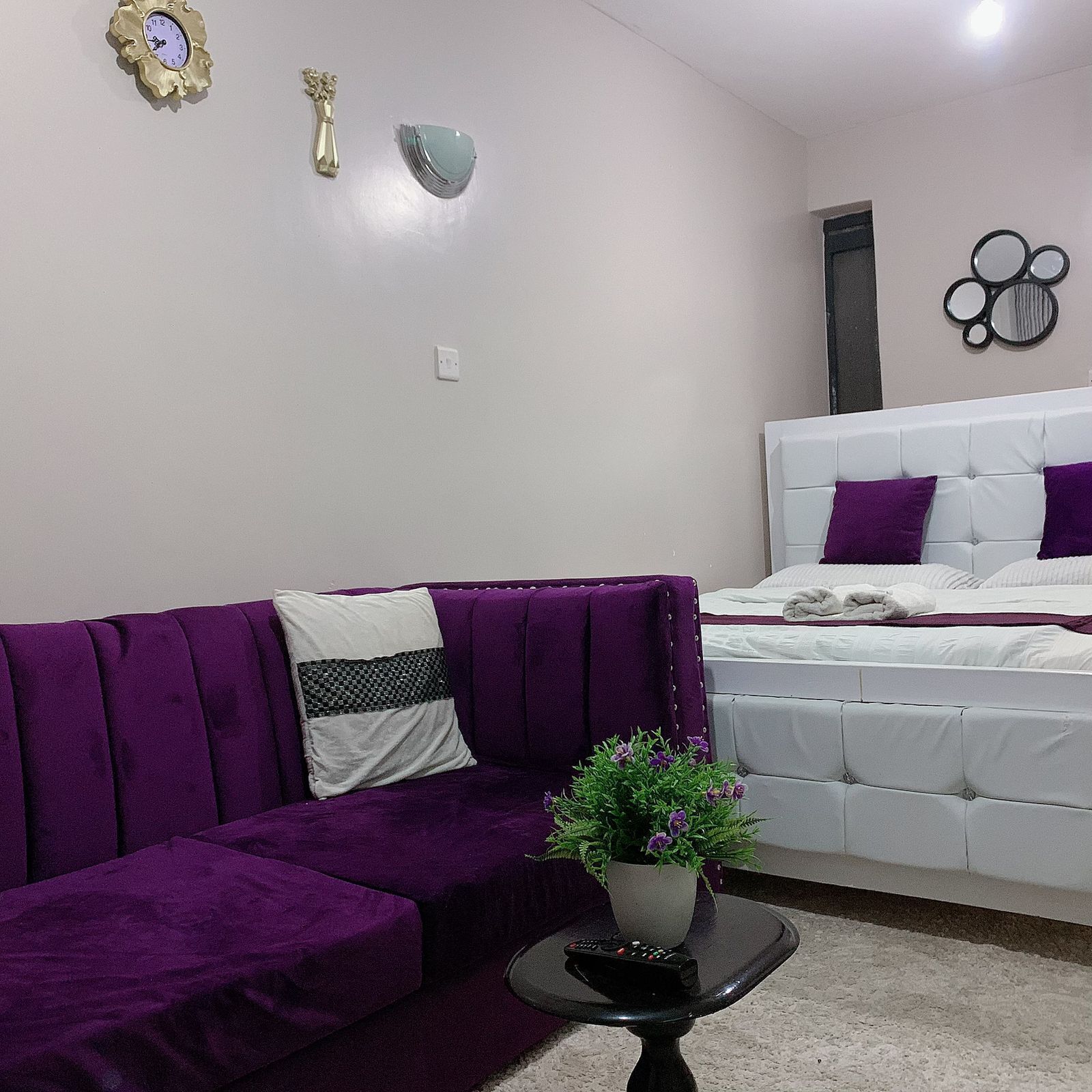 A modern bedroom with a purple sofa, a white bed with purple pillows, a small table with a plant, and wall decorations including a clock and a mirror.
