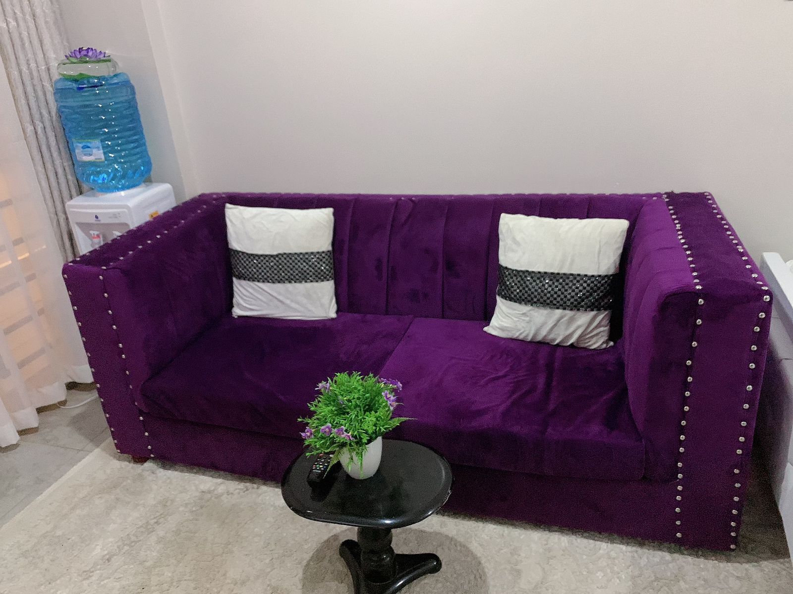 A purple sofa with two pillows, a small black table with a plant, and a water dispenser in the background.