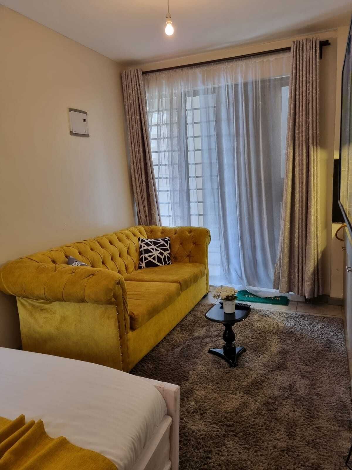 Una habitación de hotel con sofá amarillo, una mesita, cama y ventana con cortinas.