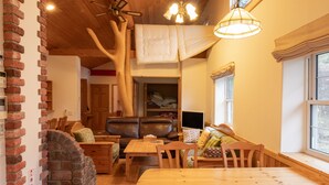 A cozy living room with a brick fireplace, wooden furniture, a dining table, and a large window with a view. - Yubari City Deer Valley Residence (Yubari)