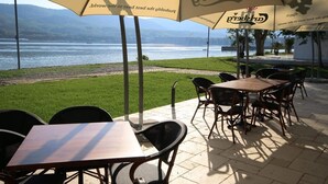 An outdoor seating area with tables and chairs under umbrellas, overlooking a body of water and greenery. - Riviera by Queens (Drobeta-Turnu Severin)