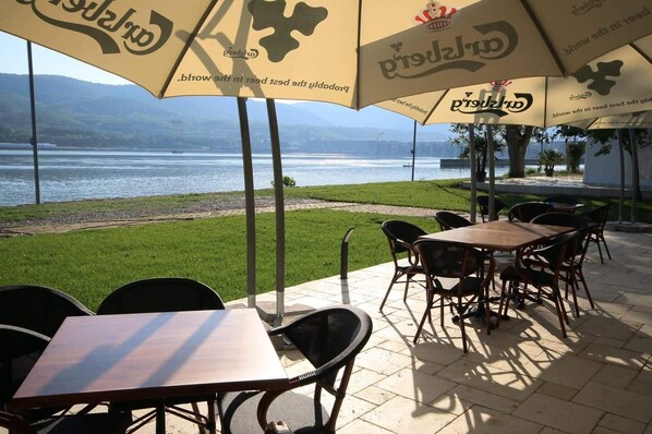 An outdoor seating area with tables and chairs under umbrellas, overlooking a body of water and greenery. - Riviera by Queens (Drobeta-Turnu Severin)