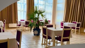 Comedor con mesas y sillas, una planta en maceta y ventanas amplias con cortinas.