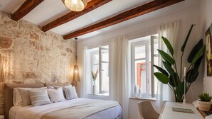 A bedroom with a stone wall, wooden beams, a bed with white bedding, a chair, a table, and a large plant. - Modern Stone Villa Lemonia w/(heated-hot tub)pool/Steam room, Old City, Rethymno (Rethymno)