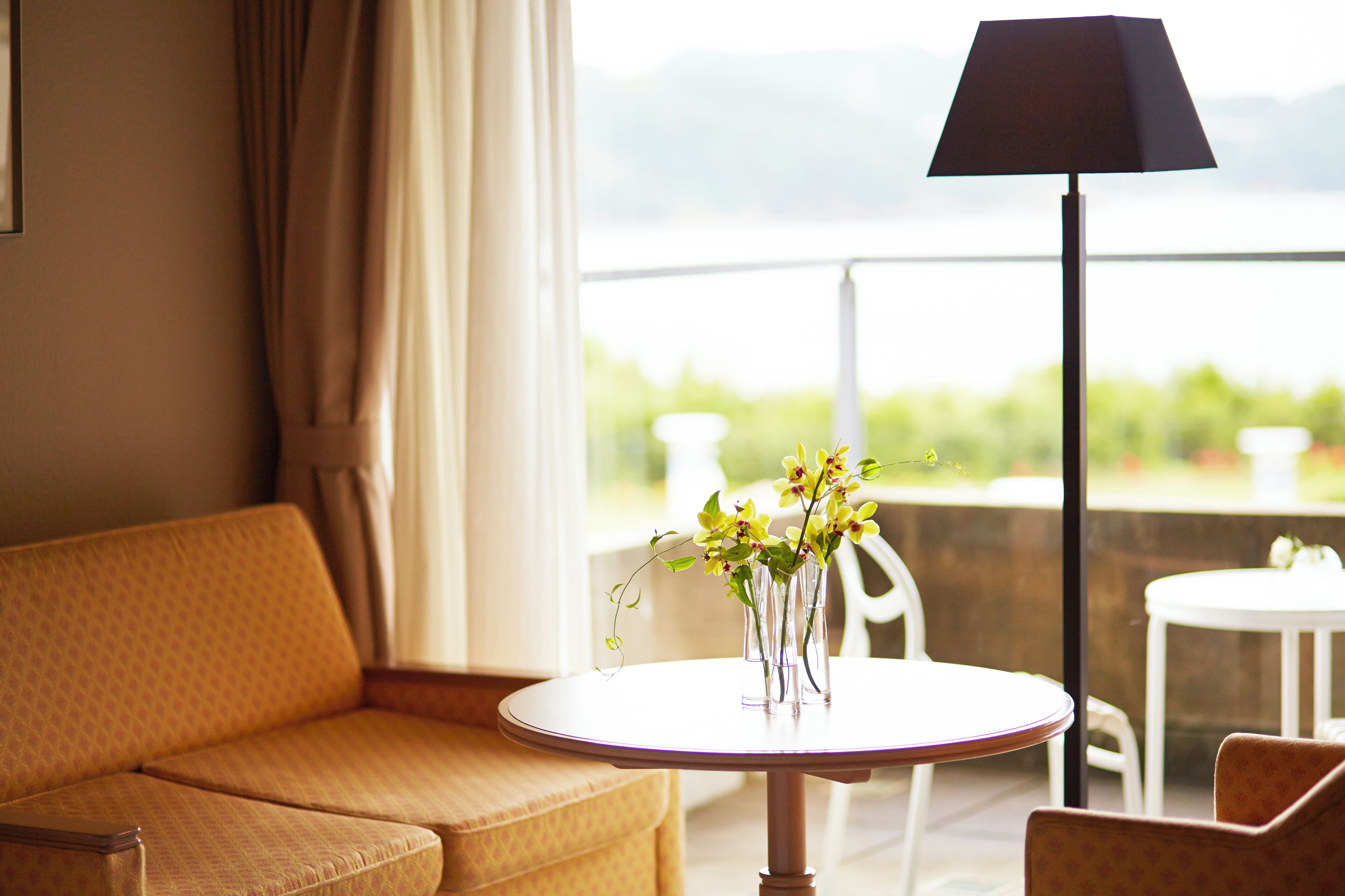 Una habitación con sofá, una pequeña mesa redonda con un florero, una lámpara de pie y vistas a la vegetación y montañas a través de una ventana.