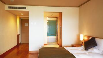 A hotel room with a bed, a nightstand, a lamp, a bathroom visible through an open door, and a wooden floor.